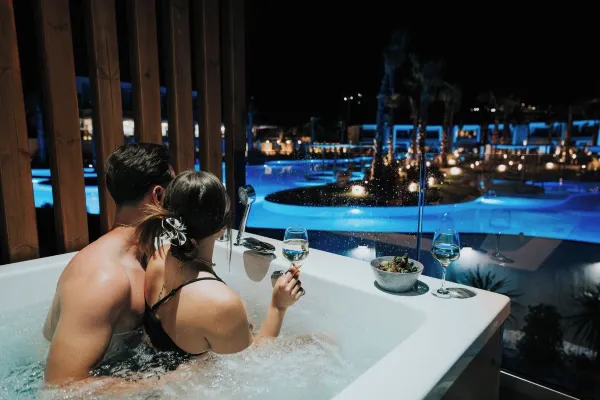 A man holding a woman in a bathtub at Anna Hotel, enjoying wine and a night view of the pool.