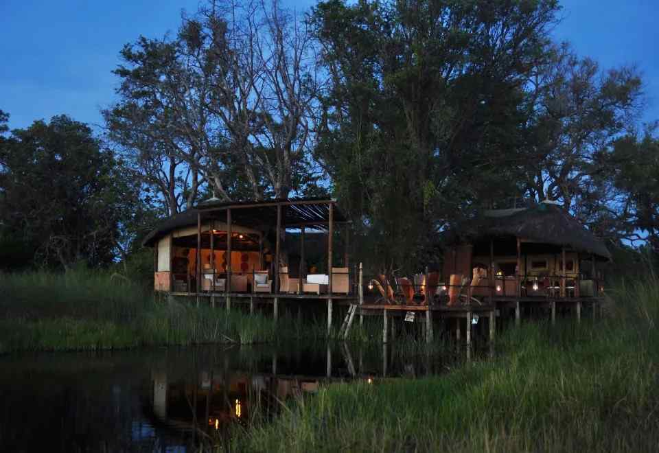 Night view of Sanctuary Baines’ Camp with reflections on water, surrounded by trees and safari-style lodges